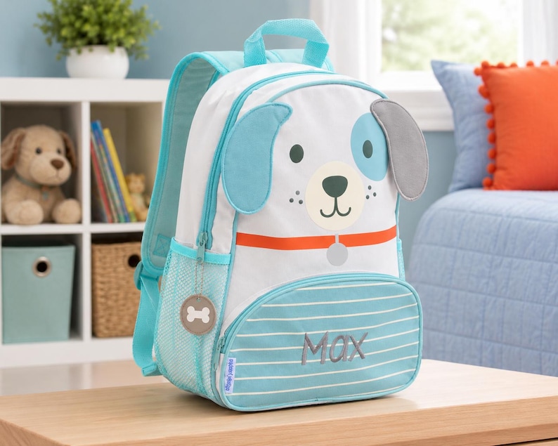 K&ouml;nnte beinhalten: Ein wei&szlig;-blauer Rucksack mit einem Hundegesicht-Design. Der Rucksack hat blaue Akzente, einen Griff und eine Vordertasche mit dem aufgestickten Namen "Max". Er hat einen Knochen-Rei&szlig;verschluss.