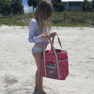 A young girl in a blue and white swimsuit stands on a sandy beach, holding a large pink bag with a rainbow pattern.
