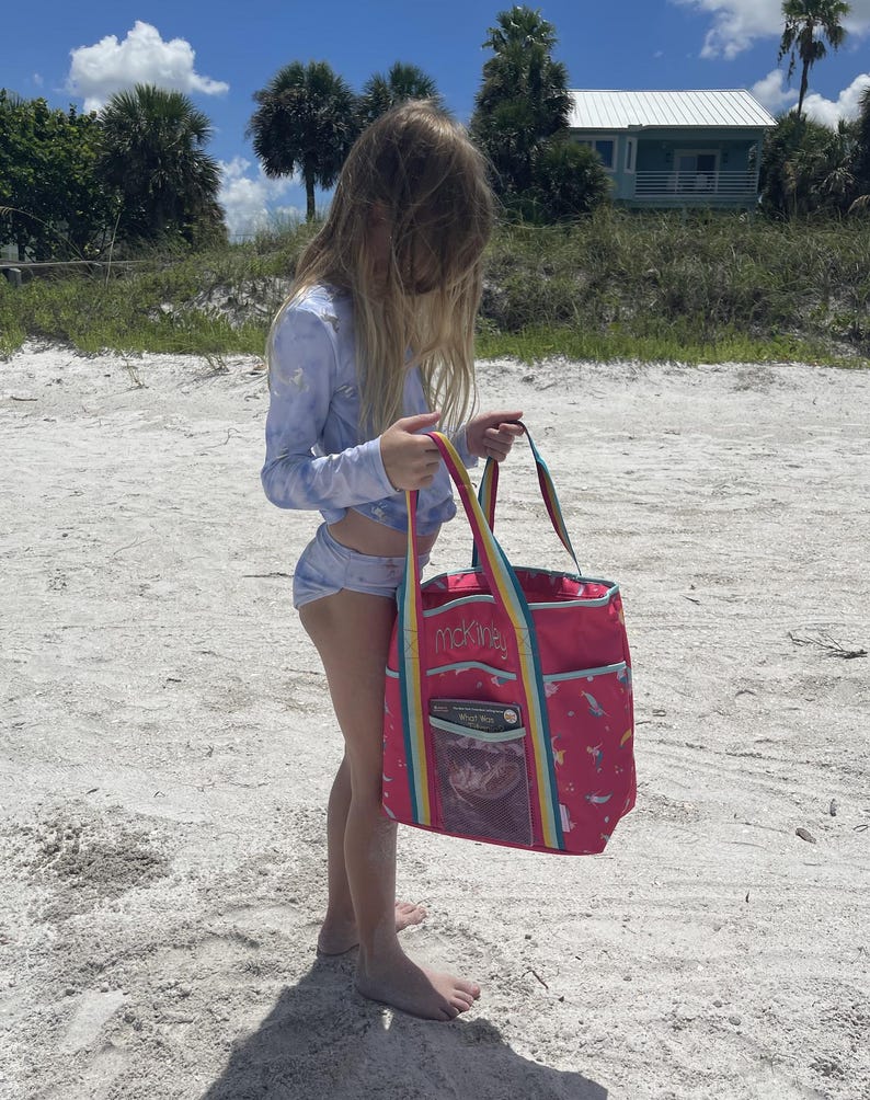 A young girl in a blue and white swimsuit stands on a sandy beach, holding a large pink bag with a rainbow pattern.