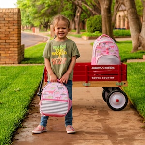 May include: A young person stands on a sidewalk, holding a pink dinosaur-themed backpack. A red Radio Flyer wagon with a matching backpack sits nearby. The person wears a green t-shirt with the text "Make the World a Better Place."