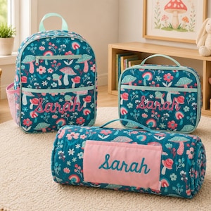 May include: A teal backpack, a rectangular lunch bag, and a rolled mat, all featuring a repeating pattern of mushrooms, flowers, and rainbows. Each item is personalized with the name Sarah embroidered in pink script on the front.