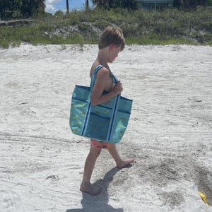 A young child wearing a blue swimsuit is walking on a sandy beach, carrying a large blue and green bag.