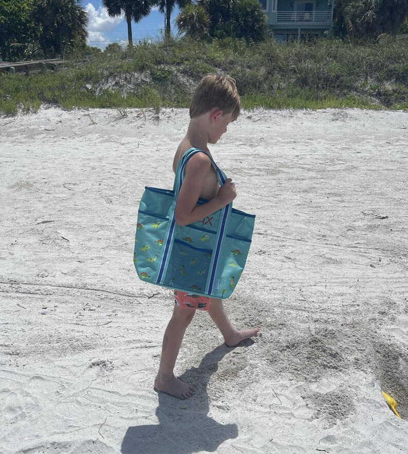 A young child wearing a blue swimsuit is walking on a sandy beach, carrying a large blue and green bag.