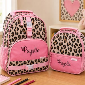 May include: A pink and leopard print backpack and matching lunch bag set. The backpack and lunch bag have a pink base with a leopard print top. The name "Payslie" is embroidered on the front of both bags. The backpack has side pockets and adjustable straps.