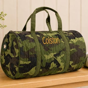 Puede incluir: Una bolsa de lona de camuflaje verde y marrón con el nombre "Colston" bordado en dorado. La bolsa tiene dos asas y un cierre de cremallera. Un pequeño dije con una estrella cuelga de la cremallera. La bolsa es acolchada.