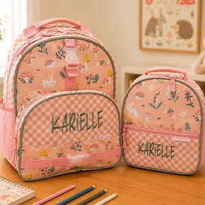 May include: A matching backpack and lunch bag set in a pink and white gingham pattern, featuring a whimsical forest print with rabbits and mushrooms. The name "KARIELLE" is embroidered on both bags. Includes side mesh pockets and a top handle.