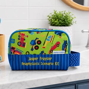 May include: A green and blue travel bag with a colorful transportation pattern. The bag features a blue striped base with the text "Jasper Fraysier Anaphylaxis Scenario Kit" in yellow. The bag is sitting on a white countertop.