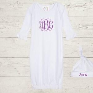 Puede incluir: Batista blanca para bebé con mangas largas y puños con volantes. La bata tiene un monograma morado en la parte delantera. Se incluye un gorro blanco anudado con el nombre "Anna" bordado en morado.