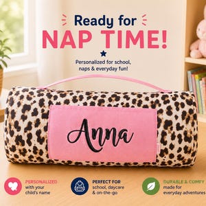 Könnte beinhalten: Eine zylindrische Schlafmatte mit Leopardenmuster, ausgestattet mit einem rosa Tragegriff und einer rechteckigen rosa Tasche, die mit dem Namen Anna in schwarzer Schreibschrift bestickt ist. Der Text Ready for NAP TIME! steht über der Matte.