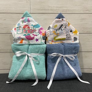 Two baby towels with personalized names on them, placed side by side on a gray surface against a wooden background.