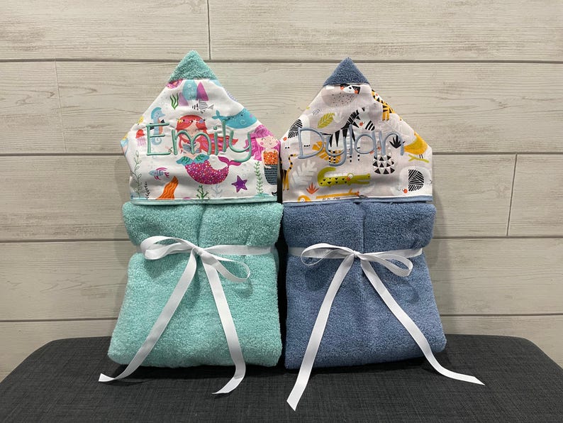 Two baby towels with personalized names on them, placed side by side on a gray surface against a wooden background.