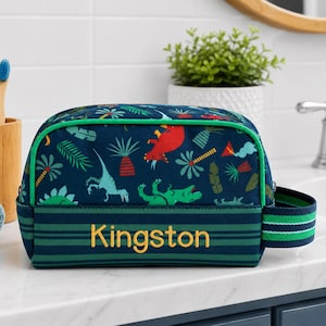 May include: A navy blue toiletry bag with a dinosaur pattern, green trim, and the name "Kingston" embroidered in gold. The bag features a green and white striped handle and a zipper closure.