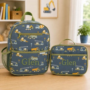Puede incluir: Una mochila y una bolsa de almuerzo a juego de color azul oscuro con un patrón repetitivo de vehículos de construcción amarillos y verdes. Ambos artículos están bordados con el nombre Glen en hilo verde y tienen ribetes y cremalleras verdes.