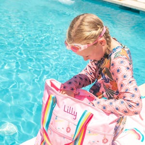 A young girl in a colorful swimsuit is sitting on a towel near a swimming pool, holding a pink tote bag with the name "Lilly" printed on it.