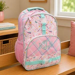 Pode incluir: Mochila rosa com estampa de unicórnio, detalhes em azul-petróleo e monograma. A mochila apresenta um design xadrez no bolso frontal e nas alças. Possui uma alça superior e um bolso lateral de malha.