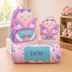 May include: A purple backpack, a matching lunch bag, and a rolled-up blanket, all featuring butterfly designs and the name Daisy embroidered in purple. The backpack and lunch bag have pink faces with antennae, while the blanket has a floral pattern.