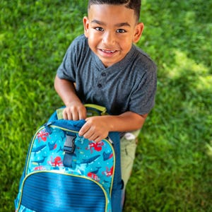 May include: A blue backpack with a marine life pattern, including whales and octopuses, is held by a smiling child. The backpack has a blue and yellow trim, a front pocket with blue stripes, and a black buckle closure. The child is wearing a gray shirt.