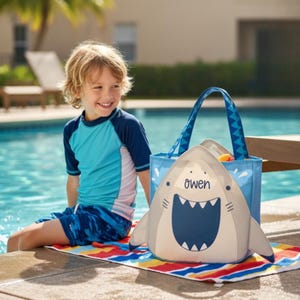 Könnte beinhalten: Eine hai-förmige Strandtasche mit dem Namen "Owen" auf einem bunten gestreiften Handtuch. Eine blaue Tragetasche befindet sich hinter der Hai-Tasche. Ein Kind in einem blau-türkisfarbenen Badeanzug sitzt am Pool.