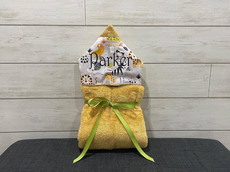 A yellow towel with a green ribbon tied around it, placed on a gray surface against a white wooden background.