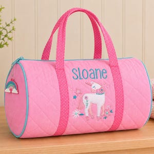 Puede incluir: Bolsa de deporte acolchada rosa con un diseño de unicornio y el nombre "Sloane" bordado en azul. La bolsa tiene asas rosas con lunares blancos, una cremallera azul y un charm de arcoíris. Una bolsa elegante y funcional.