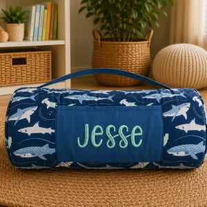 Könnte beinhalten: Eine zylindrische, marineblaue Schlafmatte mit einem sich wiederholenden Muster aus weißen und hellblauen Haien. Die Matte verfügt über eine einfarbig blaue, rechteckige Tasche auf der Vorderseite mit dem Namen Jesse in hellgrüner Schreibschrift.