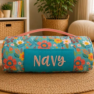 May include: A cylindrical bolster pillow featuring a vibrant teal background with a colourful floral pattern of red, orange, yellow, and white flowers. A solid teal rectangular patch on the front displays the name Navy in light pink script.