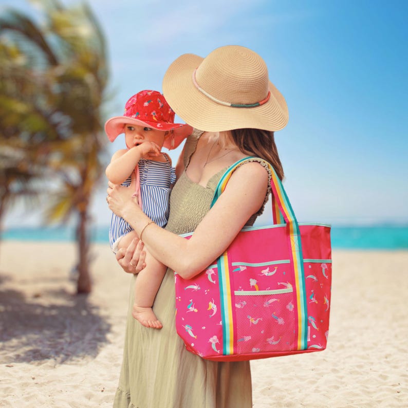 A woman in a hat holds a baby on a beach, with palm trees and a clear blue sky in the background.