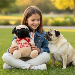 May include: A young person sits cross-legged on green grass, holding a plush pug toy with "Bella" embroidered on it. A real pug dog sits nearby, looking at the person. The plush toy has a black face and ears, with a red bow.