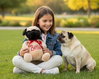 May include: A young person sits cross-legged on green grass, holding a plush pug toy with "Bella" embroidered on it. A real pug dog sits nearby, looking at the person. The plush toy has a black face and ears, with a red bow.