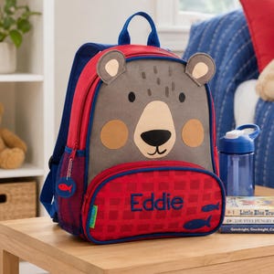 Op de afbeelding: Een rode en blauwe rugzak met een berengezichtontwerp. De rugzak heeft een rode voorzak met de naam "Eddie" erop geborduurd. Een blauwe waterfles en kinderboeken liggen op een houten tafel.