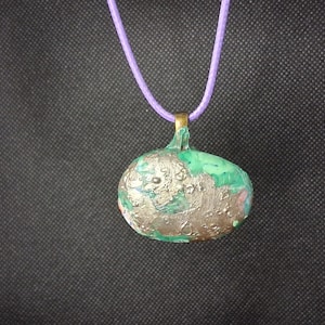 Peut inclure: Un pendentif de forme arrondie et irrégulière avec une finition marbrée verte et argentée métallique. Le pendentif est suspendu à un cordon violet clair et comprend une petite bélière en métal doré pour la fixation au collier.