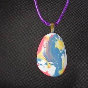 Peut inclure: Un pendentif composé d'une pierre lisse et arrondie avec une finition en peinture marbrée dans des tons de bleu, jaune, blanc et rose. La pierre est fixée à une bélière métallique et suspendue à un cordon violet vif, formant un accessoire coloré.