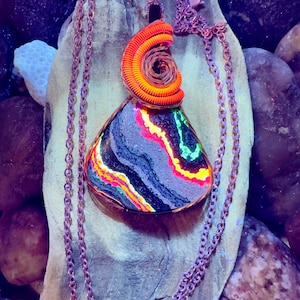 May include: A handcrafted pendant necklace featuring a colorful, geometric stone with a copper wire wrap and chain. The stone displays vibrant neon colors, including pink, yellow, and green, set against a dark background. The pendant is approximately 2 inches in length.