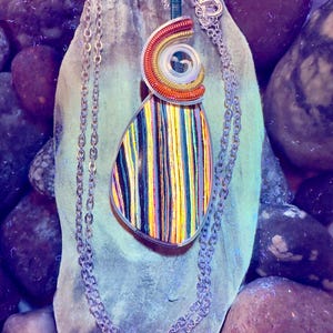 May include: A handcrafted pendant necklace with a silver chain. The pendant features a teardrop shape with colorful vertical stripes and a spiral design in shades of orange, yellow, and red. The necklace is displayed on a piece of driftwood.