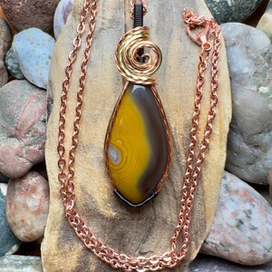 Op de afbeelding: Een handgemaakte hangerketting met een druppelvormige agaatsteen in gele en bruine tinten. De steen is bedraad met koper en goudkleurig metaal, hangend aan een koperen ketting. De ketting ligt op een stuk hout.