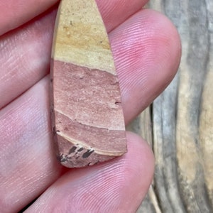 Op de afbeelding: Een kleine, driehoekige stenen hanger met een gelaagd patroon van lichtbruin, roze en wit. De steen heeft een ruwe, natuurlijke textuur.