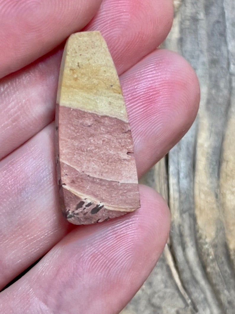 Op de afbeelding: Een kleine, driehoekige stenen hanger met een gelaagd patroon van lichtbruin, roze en wit. De steen heeft een ruwe, natuurlijke textuur.