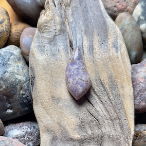 Op de afbeelding: Een paarse en witte agaat cabochon met een traanvorm. De cabochon ligt op een stuk drijfhout.