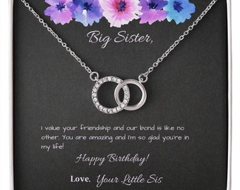 Könnte beinhalten: Eine silberne Halskette mit zwei ineinandergreifenden Kreisen, von denen einer mit klaren Kristallen besetzt ist. Die Geschenkbox trägt den Text "Big Sister, Happy Birthday! Love, Your Little Sis."