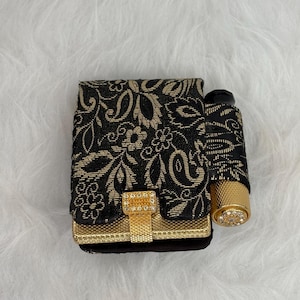May include: A rectangular black and gold case featuring a floral pattern, a gold textured clasp with embedded crystals, and an attached cylindrical gold lipstick holder with a crystal-encrusted top, all presented as a single compact accessory.