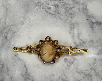 Puede incluir: Broche de camafeo dorado con el perfil de una mujer. El camafeo ovalado está rodeado de pequeñas perlas y un marco dorado decorativo. El broche tiene un diseño delicado y ornamentado.