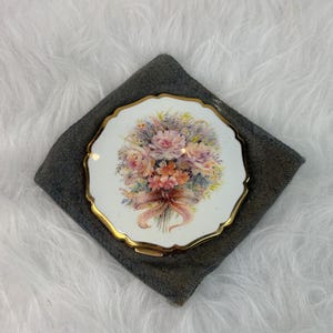 May include: A round, gold-rimmed compact mirror with a scalloped edge. The top surface is white and features a detailed, colorful illustration of a floral bouquet tied with a ribbon in shades of pink, orange, yellow, and blue, set against a white background.