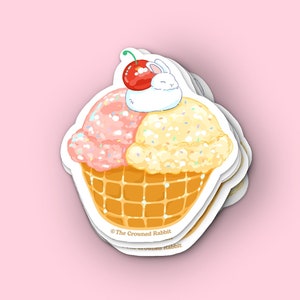 Peut inclure: Un autocollant repr&eacute;sentant un c&ocirc;ne de cr&egrave;me glac&eacute;e dessin&eacute; avec deux boules de cr&egrave;me glac&eacute;e, une rose et une jaune, surmont&eacute;es d'une cerise et d'un lapin blanc. Le c&ocirc;ne est de style gaufr&eacute; et la cr&egrave;me glac&eacute;e est d&eacute;cor&eacute;e de p&eacute;pites. L'autocollant porte le texte "@The Crowned Rabbit" en bas.
