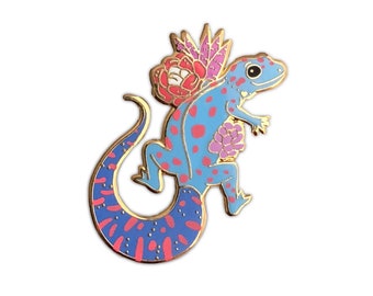 May include: A blue and pink enamel pin of a gecko with a floral design on its back. The gecko is standing on its hind legs with its tail curled around its body.