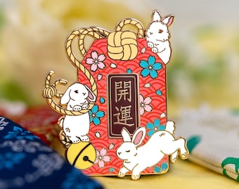 May include: A red and gold enamel pin featuring three white rabbits and a red and gold tag with the text "開運" in Japanese. The tag is decorated with cherry blossoms and a gold rope.