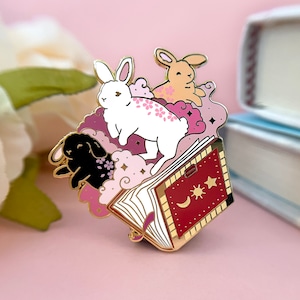 May include: Gold enamel pin featuring three rabbits sitting on a closed book with a red cover. The book has a crescent moon and sun with a star on the cover. The rabbits are white, black, and brown with pink and white clouds behind them.