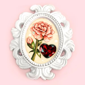 May include: A white ornate oval frame with a pink carnation and a red heart-shaped gem inside. The frame has a beaded border and intricate scrollwork.