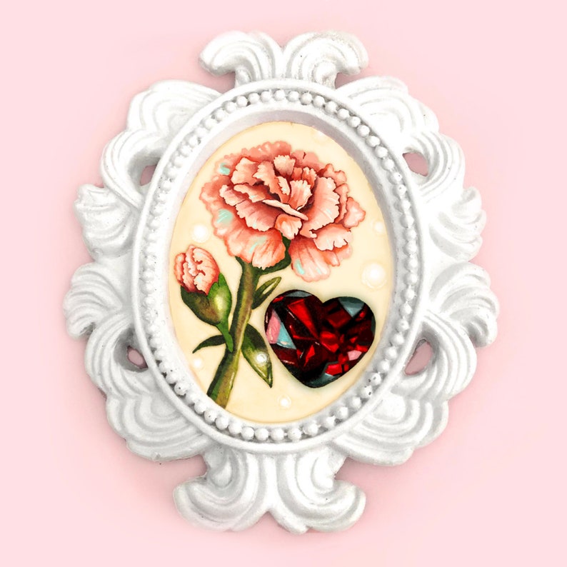 May include: A white ornate oval frame with a pink carnation and a red heart-shaped gem inside. The frame has a beaded border and intricate scrollwork.
