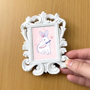 May include: A white rabbit with blue eyes holds a red knife in its paws. The rabbit is sitting on a pink background with white stars and a red heart. The image is framed in a white ornate frame.