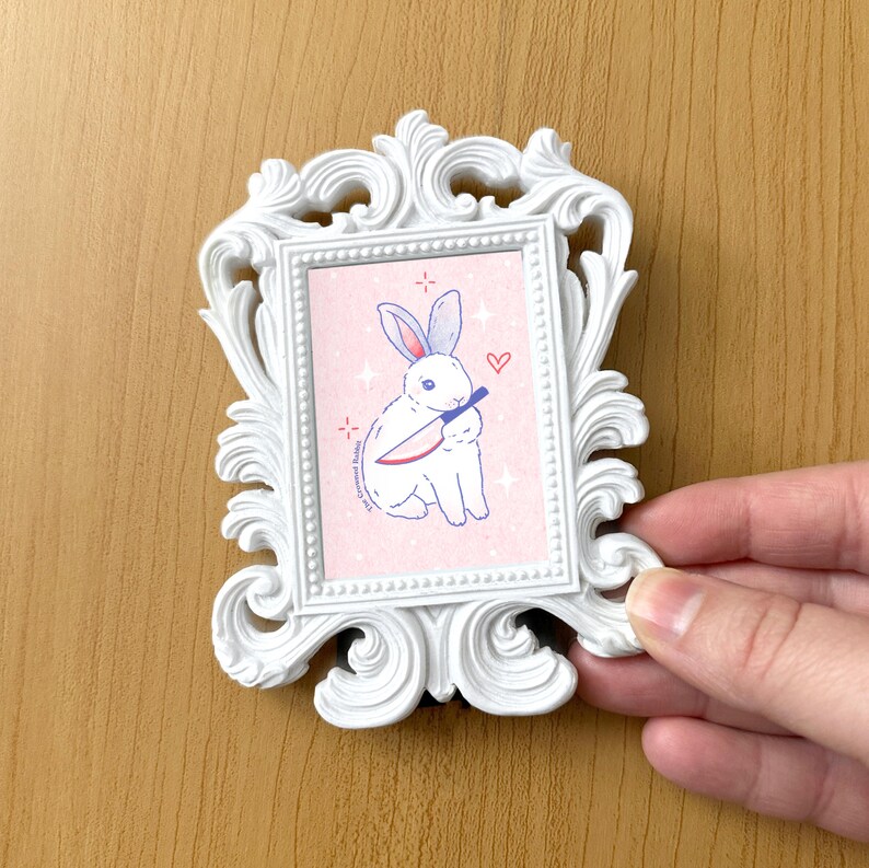 May include: A white rabbit with blue eyes holds a red knife in its paws. The rabbit is sitting on a pink background with white stars and a red heart. The image is framed in a white ornate frame.